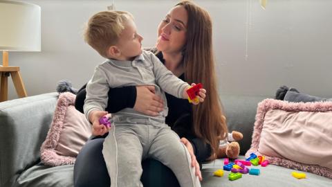 Beth and her son Fernee are sitting on a grey sofa with pink pillows. They are looking at each other. Beth has long brown hair and is wearing a black top and trousers. Fernee is a little boy wearing a grey tracksuit. He holds a coloured brick in each hand. 