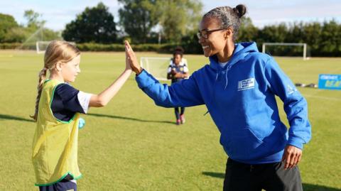 Rachel Yankey giving a girl a high five during her visit to an Oxford football training session run by Oxford United for Sport England backed campaign Play Their Way. They are smiling. Another girl can be seen behind thee. It's a sunny day.