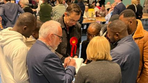 People standing around at an election count. One has a red Labour rosette.