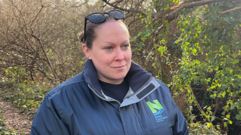Woman smiling in blue jacked with green logo on it, standing in front of trees. 