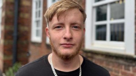 A young man with blonde hair, black tshirt and a chain around his neck, standing in front of a brick building