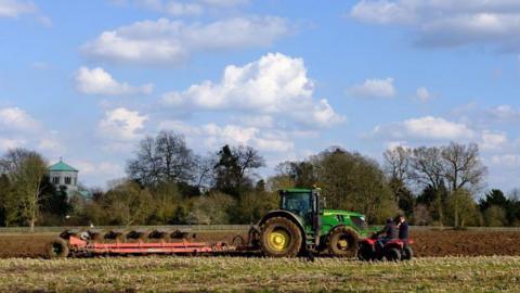 A side-on view of a green tractor ploughing a field. Two people on quad bikes can be seen next to it. 