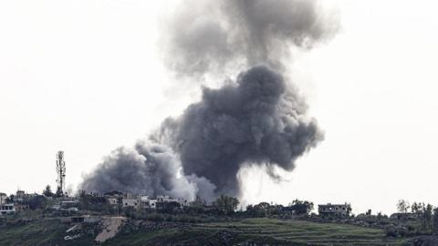 A large grey bundle of smoke rising from a village on a hillside. The smoke cloud fills most of the picture and is dramatic against the backdrop 