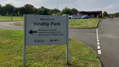A sign at Hindlip Park, which is the headquarters of West Mercia Police, points visits and staff to the entrance. It is it a road junction surrounded by areas of grass. There are road markings and buildings and cars in the background.
