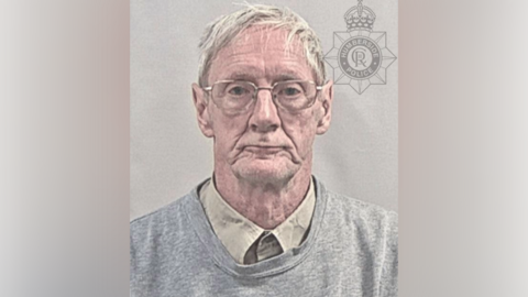 A police custody photo of Edward Sherriff. He has short blond hair and is wearing glasses, a beige shirt and a grey sweatshirt. The wall behind him is grey. 