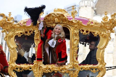 A woman in municipal robes waves from the door of an 18th century golden coach. 