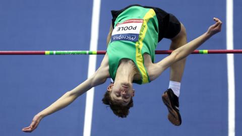 A teenager wearing a green and yellow top contorts his body in jumping over a high jump pole head first 