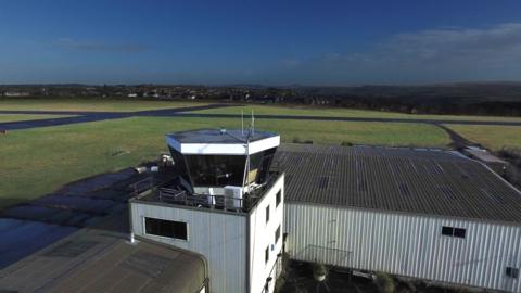 A glass air traffic control tower and a warehouse-like building. Behind it is an airport runway.