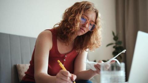 Young women looking at her finances with a computer and a glass of water in front of her and a pen and paperwork in her hands.
