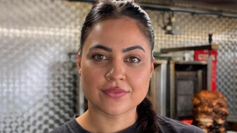 Sahar with tied-back black hair looks at the camera while inside her food van. There is an oven and a spit with roasted meat behind her.  