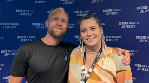 Will Crawfrod and Rachel Boss stand side-by-side, smiling for the camera in a radio studio. Will has blonde hair and a beard, and is wearing a dark t-shirt. Rachel, has dark hair with some braids and is wearing a colourful patterned top, with a lanyard around her neck displaying several buttons. Behind them is a dark blue wall with "BBC RADIO JERSEY" printed repeatedly in white.
