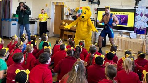 A giant costumed Pudsey bear and TV personality Gladiator Steel in front of around 20 young children with their backs to camera wearing red jumpers and Pudsey ears