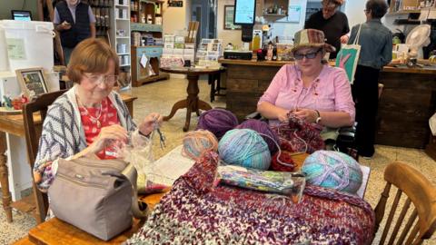 Two women, one in pink top and the other wearing red, knitting over a table in a coffee and refills shop. A man is serving a woman over the counter while a man in the background stands scrolling on his phone.