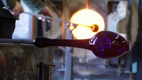 Photo inside Bristol's blue glass factory. This picture shows a blue vase being shaped on a stick in the factory. In the background you can see a man putting a a long stick into furnace.