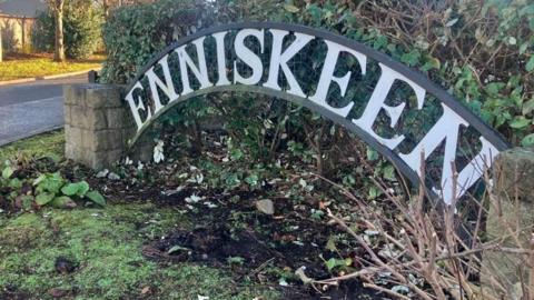 A sign displaying the name Enniskeen. It is in front of a green hedge and there are stone pillars either side