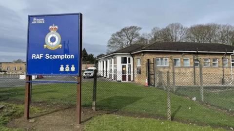 The blue RAF Scampton outside the former air base. Some of the buildings can be seen behind the fence. 