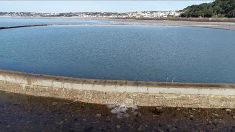 A drone shot of the Victoria bathing pool, it's a circular bathing pool with Saint Aubin's bay in the background