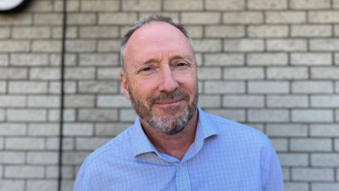 Deputy Paul Montague - A man with grey and black hair and a blue open shirt in front of a grey brick wall. 
