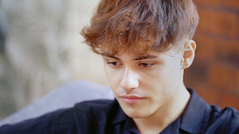 A close-up of Cai, a young man wearing a black shirt, looking pensive with his eyes cast downward