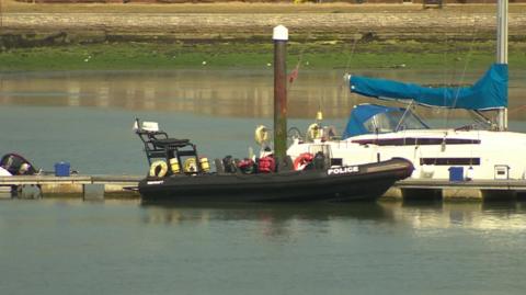 Black police boat a large, black rigid hulled inflatable boat on water alongside a pontoon with other boats in the distance.