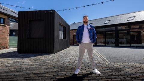 Man in blue jacket and white T-shirt stands in front of a black box on a sunny day.