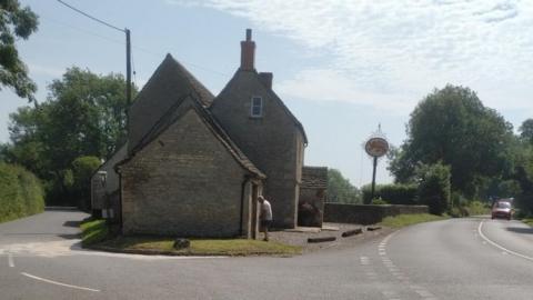 A stone-brick pub stands at the intersection of two roads. There are lots of trees and hedgerows around, and it is a sunny day with light cloud cover. 