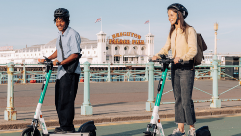 Two people on e-scooters on a road. They are wearing helmets. A pier can be seen in the background.