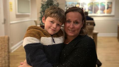 A boy with brown hair, a woolly brown, white and black top sitting on the lap of a woman with a dark top and brown hair, who is cuddling him