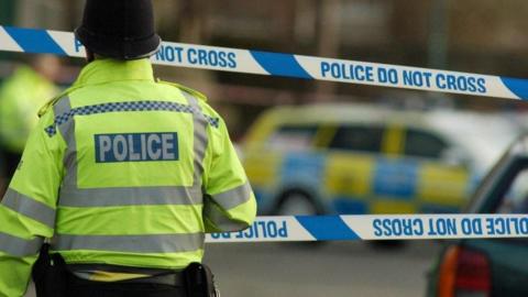 A police man on the left wearing a yellow hi-vis jacket and a police hat. He is approaching two lines of blue 'do not cross' police tape which is cordoning off an area. In the distance you can see a blurred out police car parked on the street.