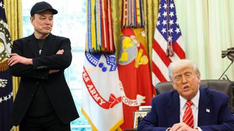 Elon Musk and Donald Trump in the oval office. Both men are wearing dark-coloured suits. Flags can be seen in the background.