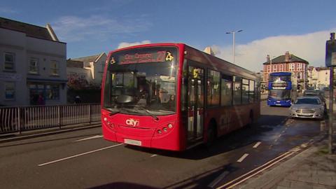 Single decker red bus on a road on a street driving towards the left of the camera passing parked cars and with a double decker bus following.