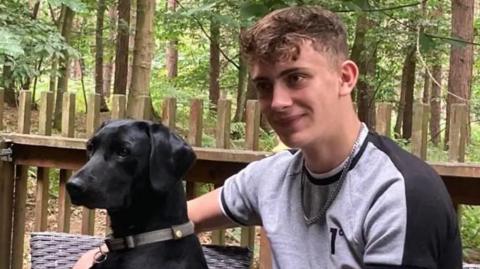 Cory Chilvers with light brown hair, curly on top, wearing a grey and black T-shirt and silver neck chain, with his arm around a black Labrador, was sitting on outdoor furniture with woodland in the background.