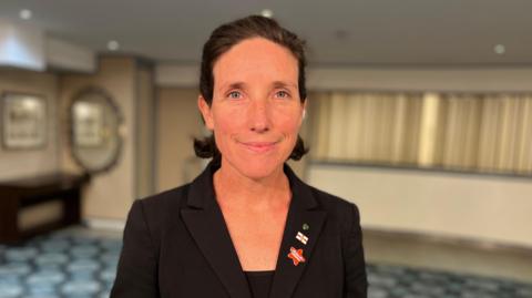 Lindsay de Sausmarez looking at the camera, she is a woman with close black hair, wearing a black jacket with three pin badges on the lapel. 