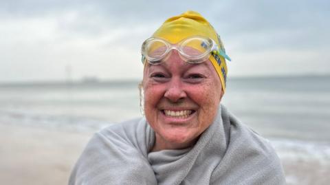 Helen Jenkins smiles into the camera after a swim, wrapped in a towel and wearing a yellow swimming hat, with goggles pushed up on the top of her head.