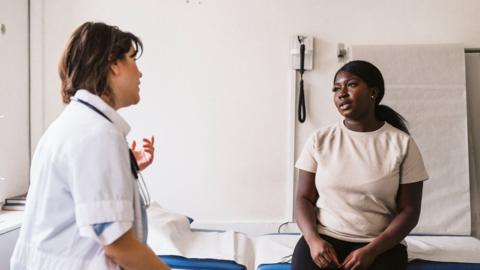 A young female sits on a blue bed at a GP's office. She is talking to a female nurse or doctor and both have a serious but neutral expression.