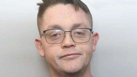 A man with brown hair shaved at the sides, and wearing thin-rimmed glasses, looks at the camera against a white background.