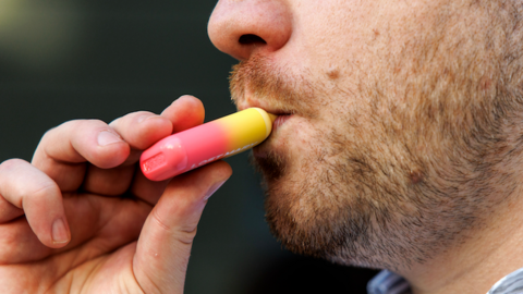 A man with stubble using a disposable lost mary vape, which is an ombre pink and yellow square shaped.