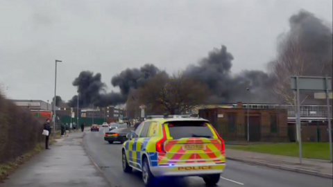 A police car drives to the scene of the factory fire with black smoke in the distance