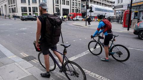 Two cyclists wait at a junction in central Londons. Cars, buses, pedestrians and a red telephone box are visible in the background.