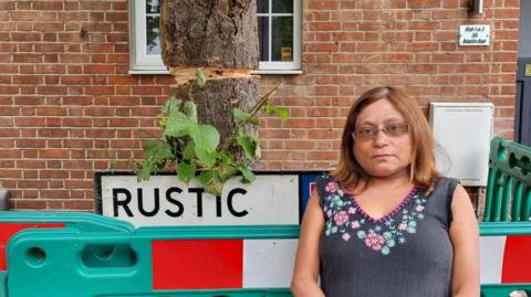 A lady with shoulder-length brown hair looks into the camera, wearing a blue top with pink and green flowers. She is stood by a tree, that has been cordoned off, because it's trunk has been partially sawed. It is behind a road sign that is partially covered, with the words RUSTIC. 