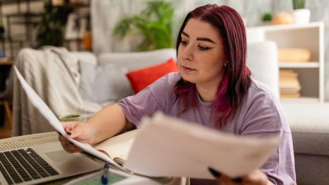 A young woman with purple hair and wearing a mauve top looks at a bill while sitting in at a laptop