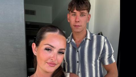 Selfie photo by Kyra Farnum with Sebastian Pixley who is standing behind her. She has long brown tied back hair and is wearing make-up. He has short brown hair and is wearing a short-sleeved short with grey and white zigzag patterns down it.
