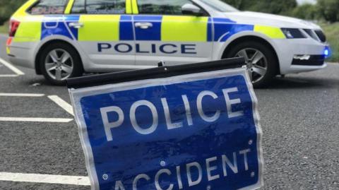 A blue and white police incident sign stands on the road, with a yellow and blue police vehicle parked in the background