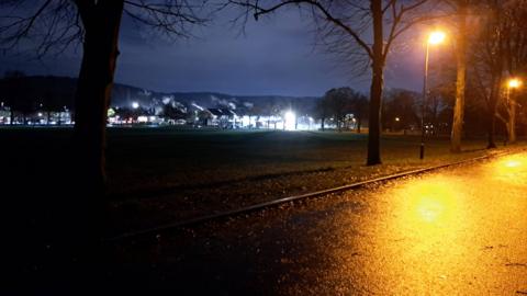 A view of a park at night shows a yellow glow from a street lamp while the rest of the park is in complete darkness with some house lights in the distance