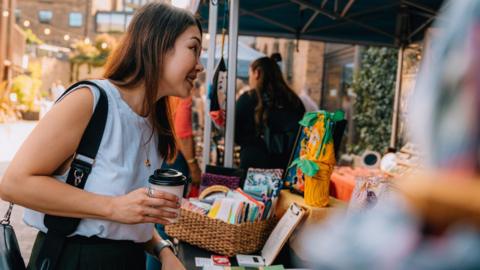 A woman with long brown hair is holding a coffee cup and is looking at a market stall with books for sale.