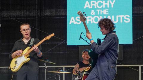 Three young male musicians perform on stage at Lincoln Live. Two guitarist are wearing navy blue boiler suits and the drummer is dressed in a black T-shirt. The band's name "Archy and the Astronauts" is on a light blue screen in the background.