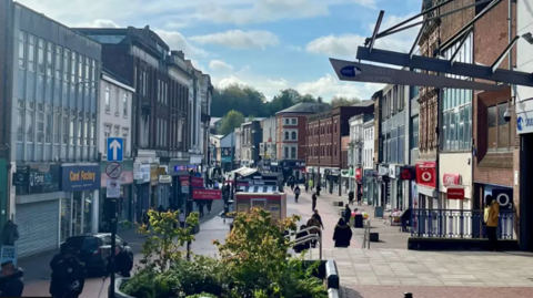 Walsall high street, with shops of different colours lining the pedestrianised route, which shows shoppers in the distance, with a flowerbed in the foreground and a car parked on the left. 