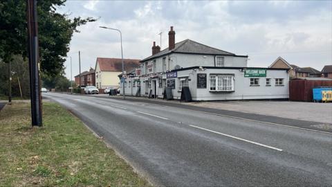 A road with a grass verge on one side and a pub on the other. There are houses beyond the pub and a parked car. There is another car on the road in the distance.