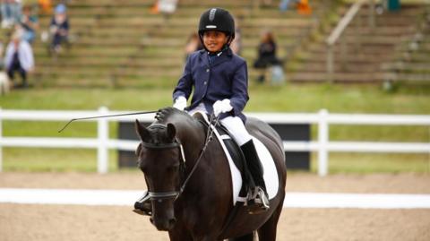 Govind in a navy suit jacket, white trousers, black boots and black riding hat. He sits on top of a dark brown horse in a riding competition ring.
