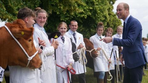 Prince William wearing a navy suit standing outside in a field at the Royal Norfolk Show pointing at a brown cow.  A group of people wearing white lab coats over shirts and ties line up next to the cow.